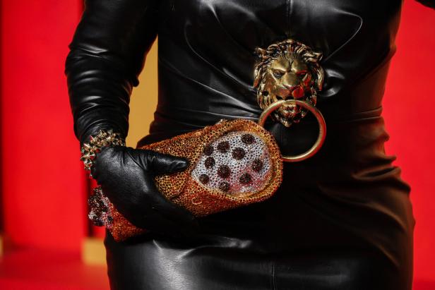 Eine Person in einem schwarzen Lederkleid hält eine verzierte Handtasche mit einem Löwenkopf-Detail.
