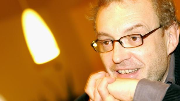 Ein lächelnder Mann mit Brille stützt sein Kinn auf seine Hände.