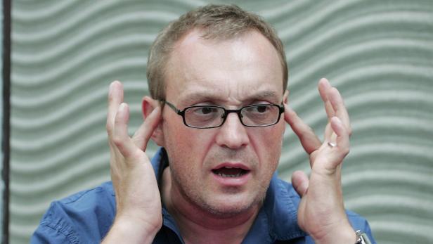 Ein Mann mit Brille gestikuliert mit beiden Händen vor einem gemusterten Hintergrund.