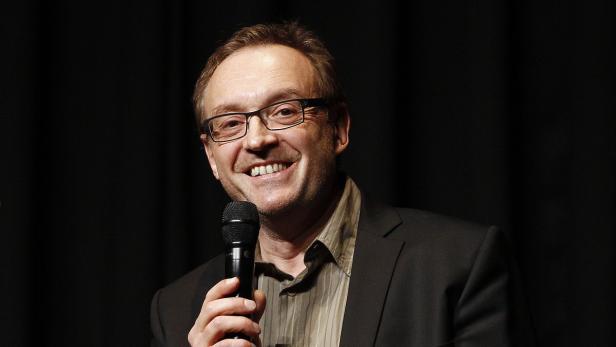 Ein Mann mit Brille lächelt und hält ein Mikrofon in der Hand.