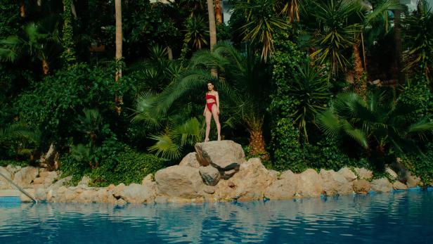 Eine Frau in rotem Bikini steht auf einem Felsen am Pool, umgeben von tropischer Vegetation.