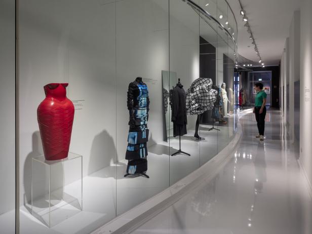 Eine Frau betrachtet in einem Museum eine Ausstellung von Mode und Kunstobjekten hinter Glas.