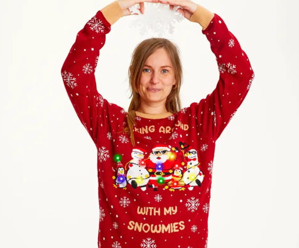 Eine Frau mit einem roten Weihnachtspullover mit Schneemann-Motiv hält eine Schneeflocke hoch.