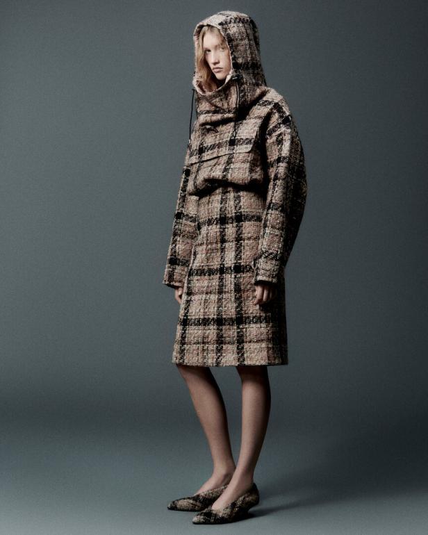 Eine Frau in einem karierten Tweed-Hoodie und Rock steht vor einem grauen Hintergrund.