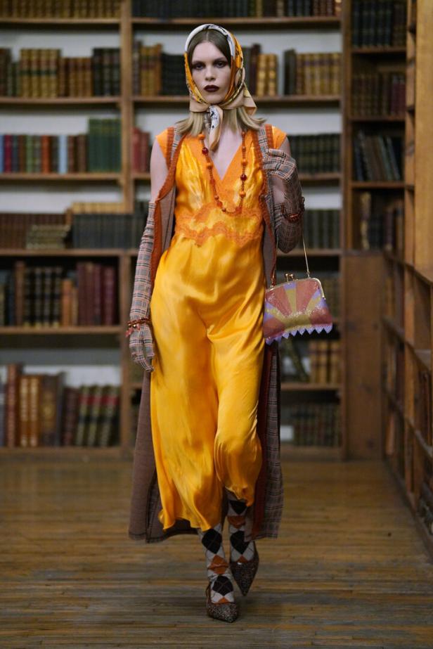 Eine Frau in einem orangefarbenen Kleid mit Kopftuch und Handtasche geht durch eine Bibliothek.