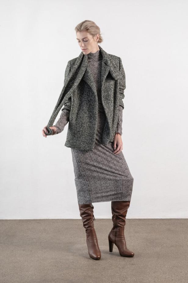 Eine Frau in einem Kleid mit Fischgrätenmuster, einem Blazer und braunen Stiefeln.