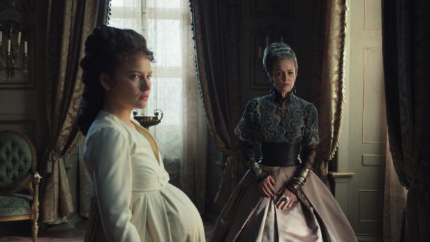 Eine schwangere Frau und eine ältere Dame in historischer Kleidung stehen in einem opulenten Raum.