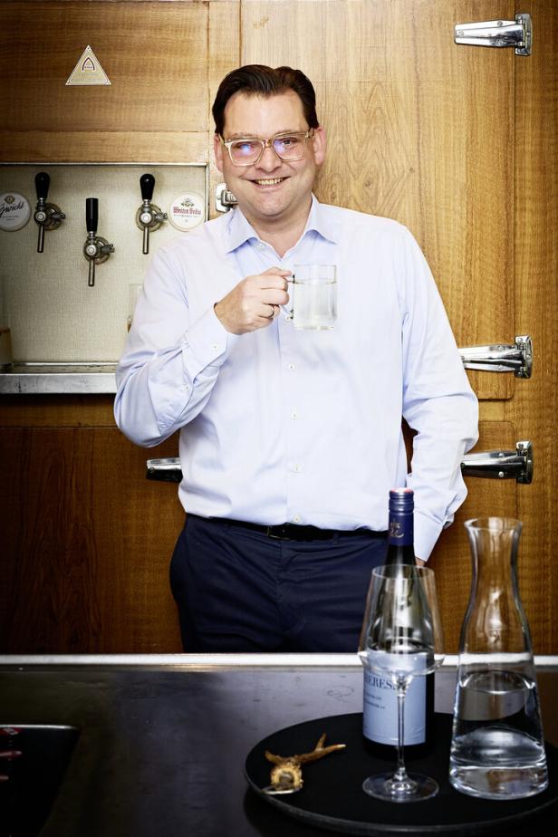 Ein Mann mit Brille hält ein Glas Wasser vor einer Holzwand mit Zapfhähnen.