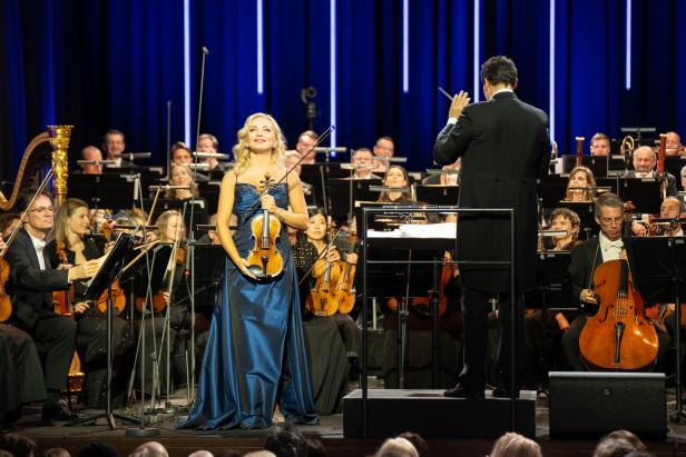 Eine blonde Frau in einem blau-schillernden Kleid mit einer Geige in der Hand auf einer Bühne vor einem Orchester