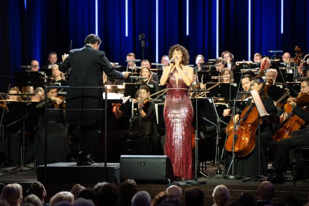 Eine Frau in einem schillernd-pinken Kleid steht auf einer Bühne mit einem Orchester und singt.