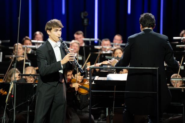 Ein junger Mann im Wrack spielt Klarinette auf einer Bühne vor einem Orchester.