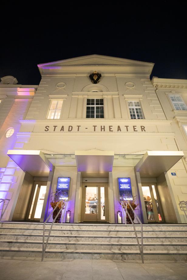 Der Eingang zum Stadttheater bei Nacht, beleuchtet und einladend, mit Menschen, die sich auf den Abend vorbereiten.