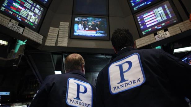 Zwei Börsenmakler mit „Pandora“-Jacken stehen vor Bildschirmen mit Finanzdaten.