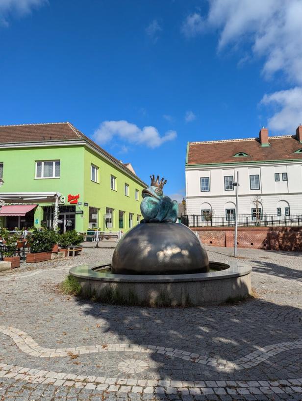 Ein Frosch mit Krone sitzt auf einem Brunnen auf einem gepflasterten Platz.