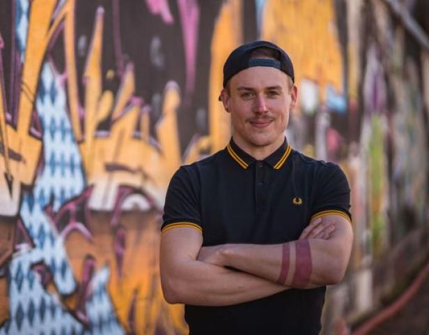 Ein Mann mit Schnurrbart und Tätowierung steht vor einer Graffitiwand.