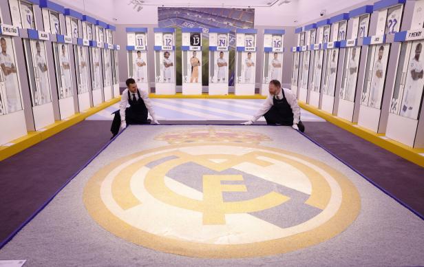 Zwei Männer rollen einen Teppich mit dem Logo von Real Madrid in einem Raum mit Spielerporträts aus.