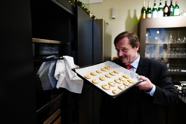 Werner Gruber lächelt vorm Backrohr und hält das Blech mit den Kipferln in die Kamera.