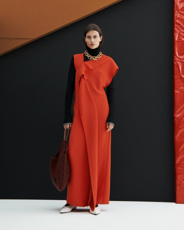 Eine Frau in einem orangefarbenen Kleid mit Rollkragenpullover und einer goldenen Halskette posiert mit einer braunen Tasche.