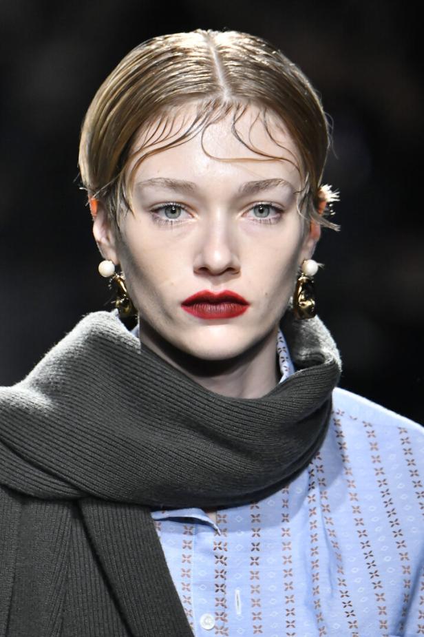Ein Model mit zurückgegeltem Haar, dunklem Lippenstift und auffälligen Ohrringen trägt einen grauen Schal.