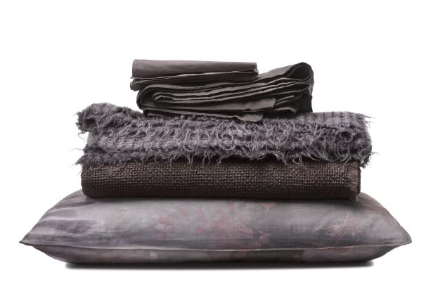 Ein Stapel aus Kissen, Decken und Bettwäsche in verschiedenen Grautönen.