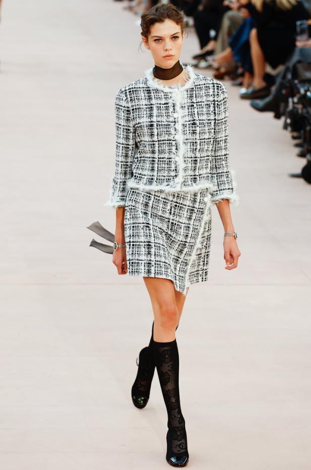 Ein Model präsentiert ein schwarz-weißes Tweed-Kostüm mit kniehohen Socken auf einem Laufsteg.