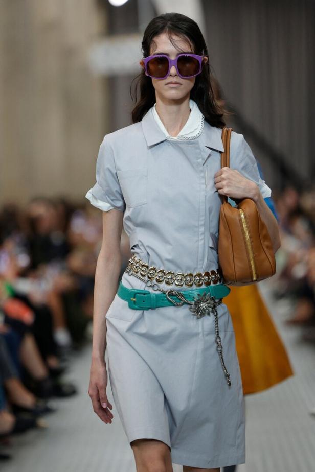 Eine Frau mit lila Sonnenbrille trägt ein hellblaues Kleid mit Gürtel und eine braune Handtasche.