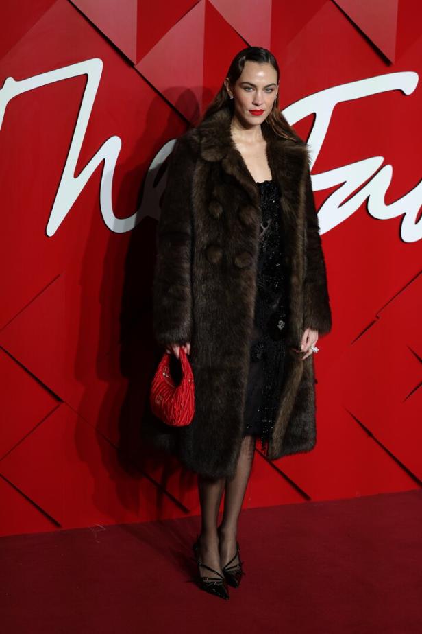 Alexa Chung trägt einen braunen Pelzmantel und eine rote Handtasche vor einem roten Hintergrund.