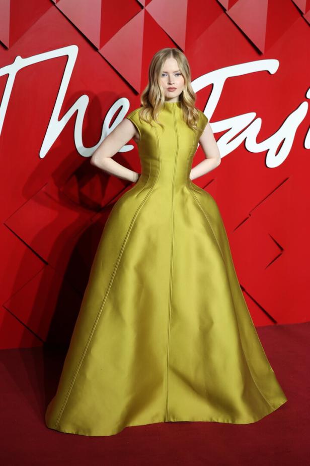 Eine Frau in einem gelben Kleid posiert vor einem roten Hintergrund.