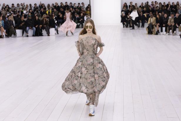 Ein Model mit Sonnenbrille präsentiert ein geblümtes Kleid auf dem Laufsteg vor Publikum.