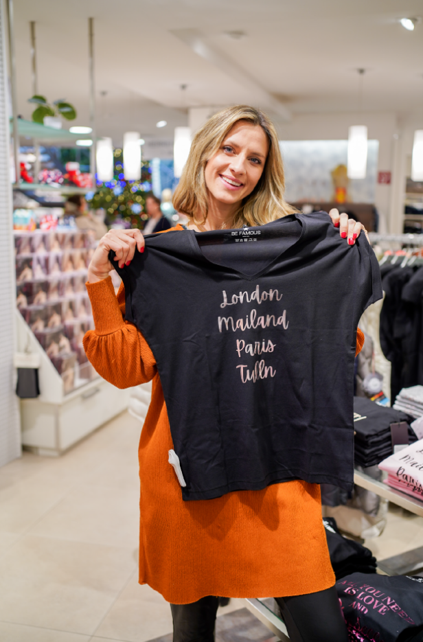 Eine Frau in einem orangen Kleid hält ein T-Shirt nach oben, auf dem "London, Mailand, Paris, Tulln" steht.