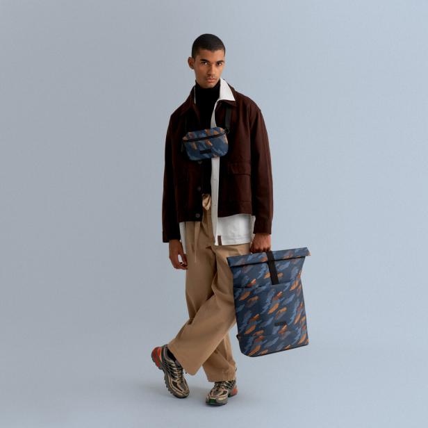 Ein Mann posiert mit einer Umhängetasche und einem Rucksack mit blauem Muster.