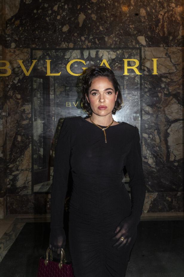 Eine Frau in einem schwarzen Kleid und mit einer Halskette steht vor einem Bulgari-Schriftzug.