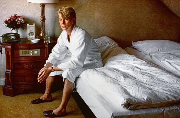 David Bowie sitzt in einem weißen Bademantel auf einem Hotelbett.