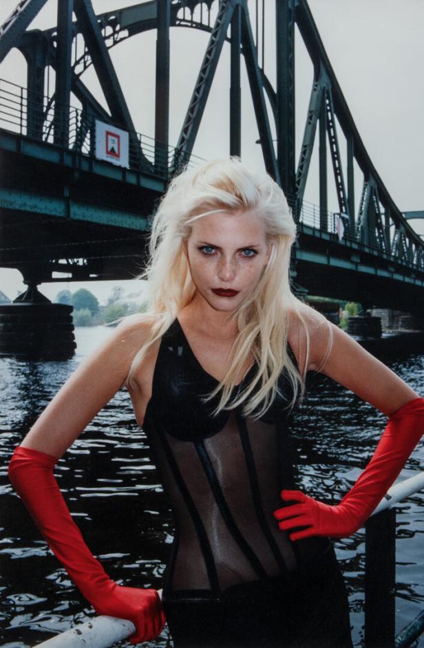 Eine Frau mit blonden Haaren und roten Handschuhen posiert vor einer Brücke am Wasser.