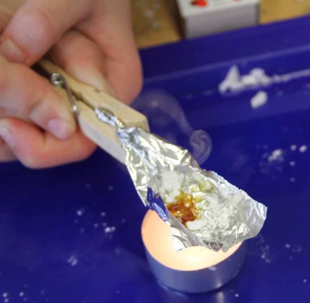 Eine Person hält mit einer Wäscheklammer Alufolie über eine Kerze, aus der Rauch aufsteigt.