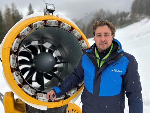 Ein Mann steht neben einer Schneekanone auf einer Skipiste.