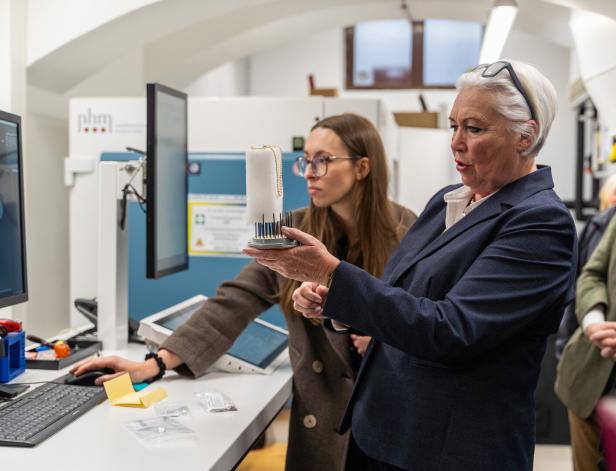 Zwei Frauen vor Monitoren und einem CT-Gerät. Eine hält einen Schaumstoff hoch, auf dem eine Perlenkette liegt.