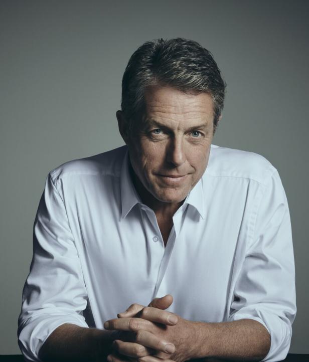 Porträt von Hugh Grant in einem weißen Hemd, die Hände gefaltet.