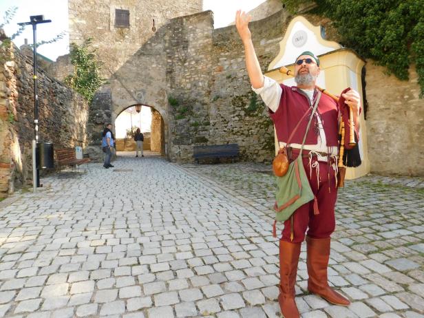 Ein Mann in historischer Kleidung steht mit einer Art Dudelsack in einer Burganlage.