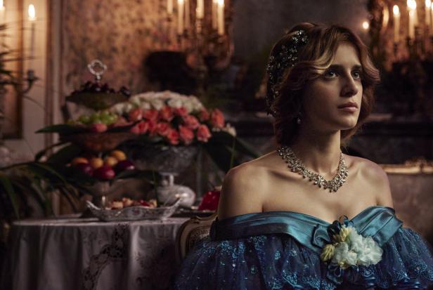 Eine junge Frau im Ballkleid mit Diadem und Halskette vor einem opulenten Tisch mit Kerzen.
