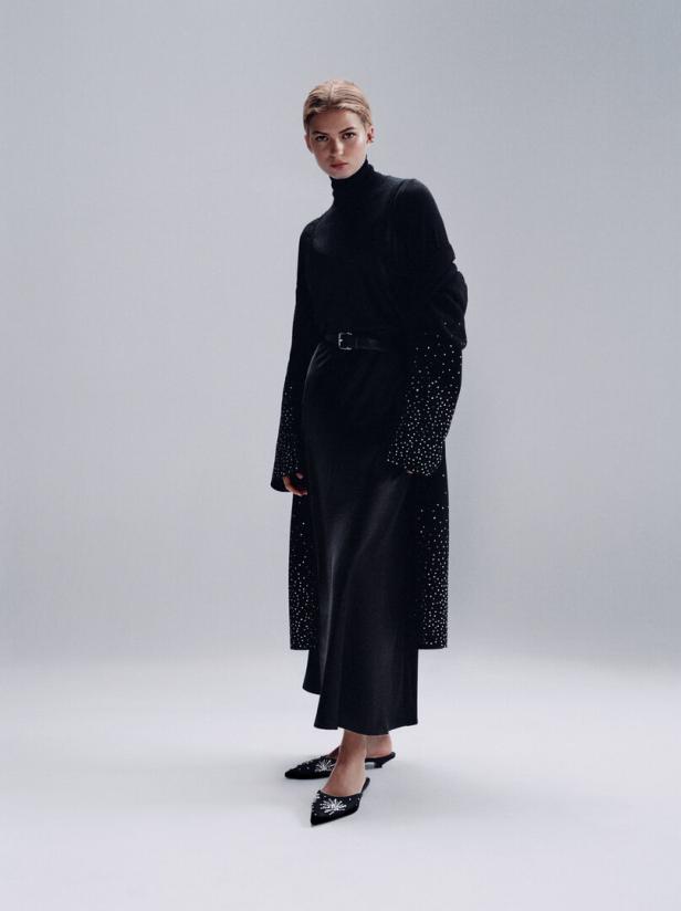 Eine Frau in einem schwarzen Rollkragenpullover, einem langen Cardigan mit Strass und einem Rock, abgerundet mit passenden Schuhen.