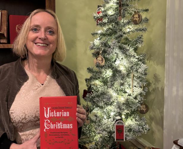 Eine Frau hält ein Buch mit dem Titel „Victorian Christmas“ vor einem Weihnachtsbaum mit Charles-Dickens-Ornamenten.