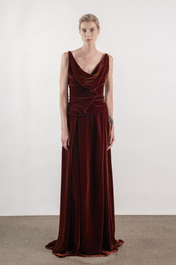 Eine Frau trägt ein bodenlanges, rostbraunes Samtkleid mit Wasserfallausschnitt.