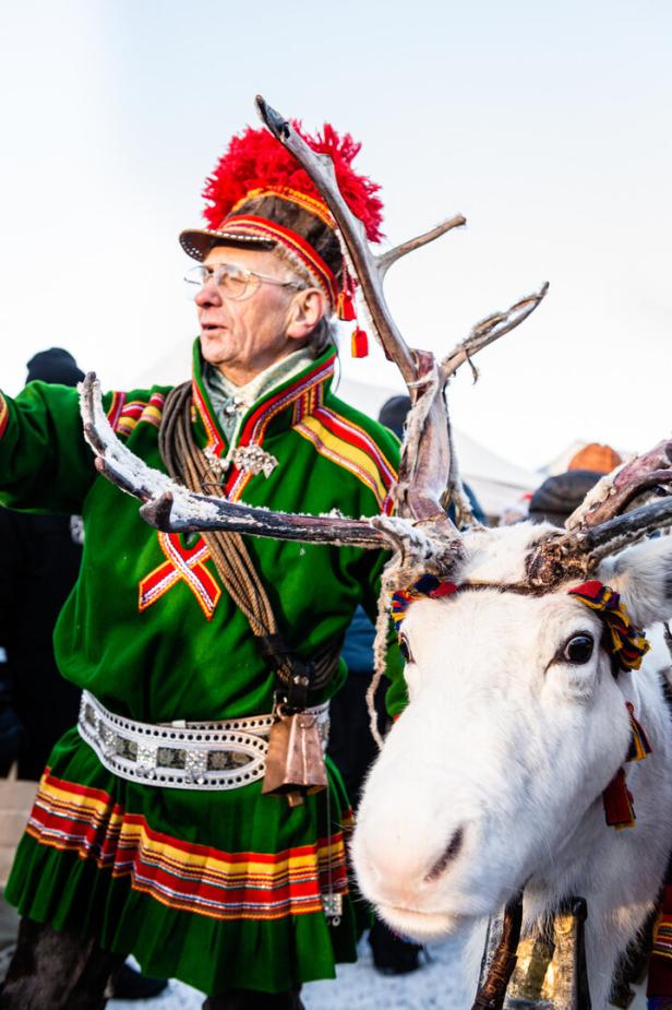 Ein Mann in traditioneller samischer Kleidung steht neben einem weißen Rentier.