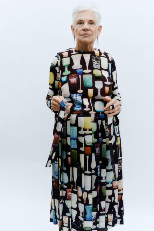 Eine ältere Frau mit grauen Haaren trägt ein Kleid mit einem Muster aus bunten Gläsern.