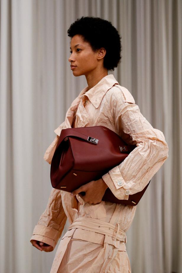 Eine Frau mit Afro trägt einen hellrosa Trenchcoat und eine große, kastanienbraune Handtasche.