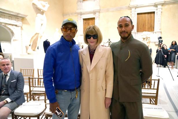 Pharrell Williams, Anna Wintour und Lewis Hamilton posieren zusammen in einem Raum mit Kunstwerken.