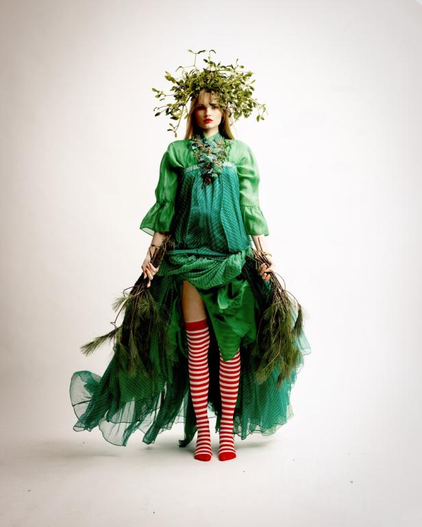 Eine Frau in einem grünen Kleid und einem Blätterkranz hält Äste in den Händen.