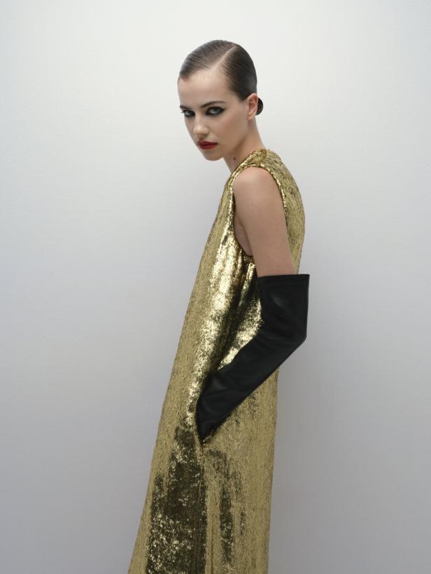 Eine Frau mit zurückgekämmtem Haar trägt ein goldenes Paillettenkleid und lange schwarze Handschuhe.
