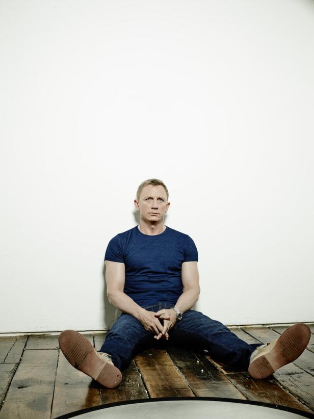 Daniel Craig sitzt in Jeans und T-Shirt vor einer weißen Wand.